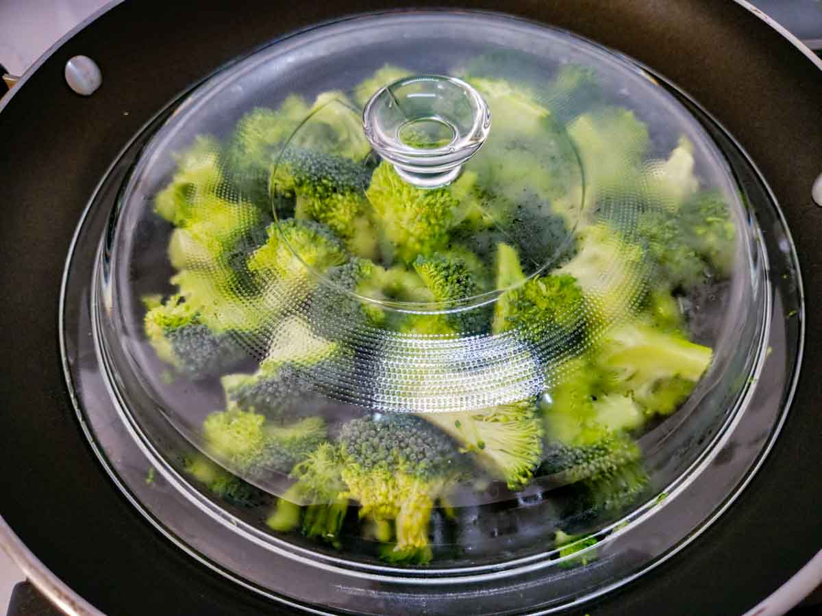 broccoli steaming under a lid in a frying pan.