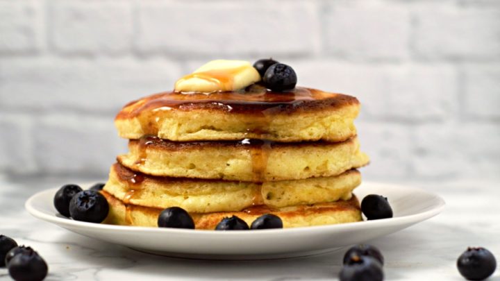a stack of 4 fluffy pancakes topped with blueberries, butter, and syrup on a plate.