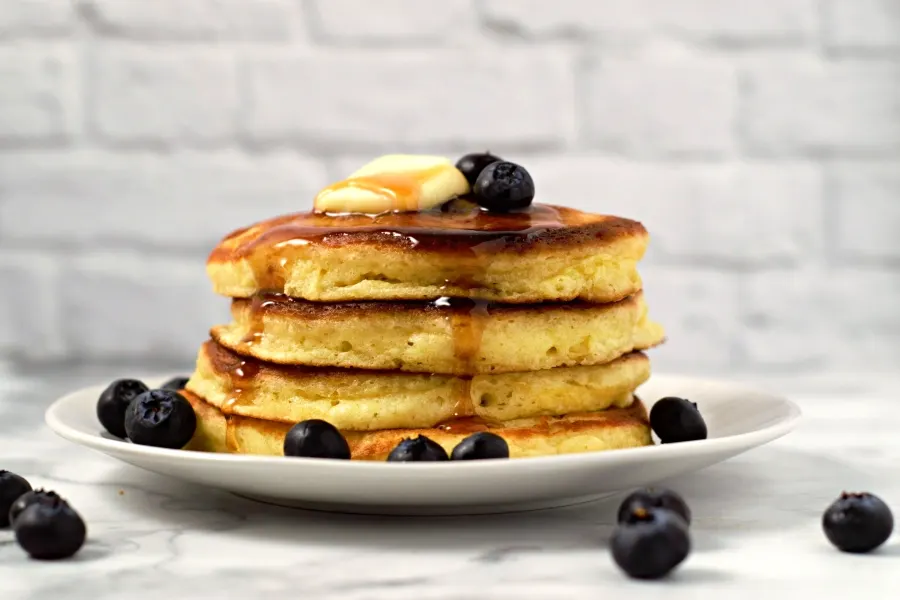 a stack of 4 fluffy pancakes topped with blueberries, butter, and syrup on a plate.