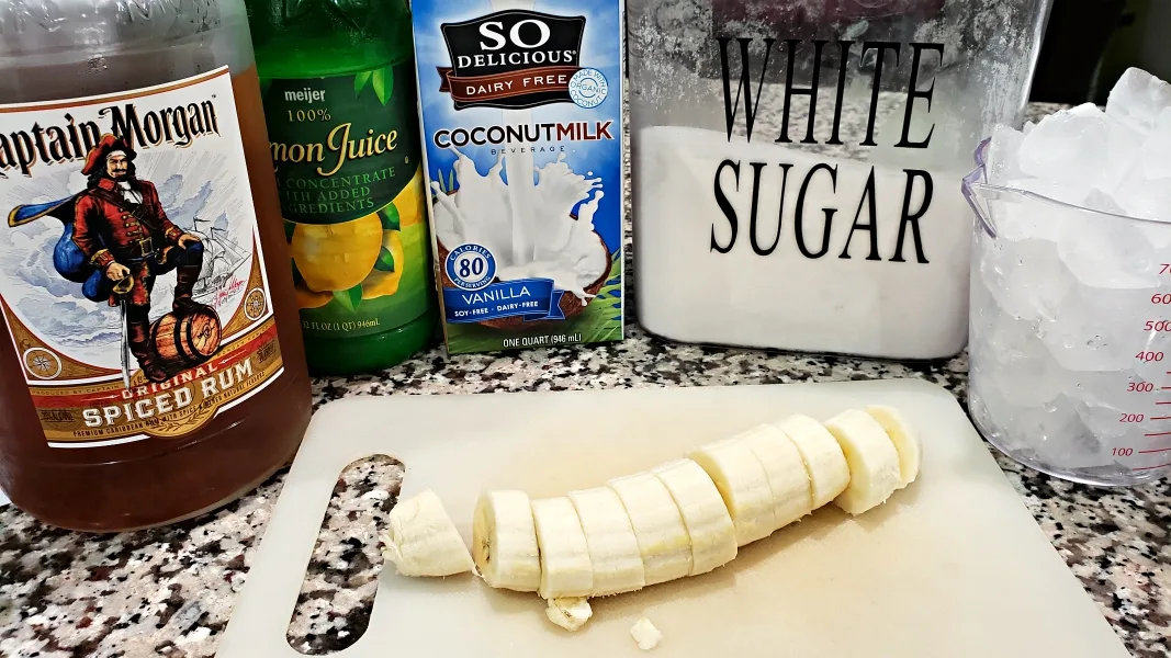 a sliced banana on a cutting board, a bottle of spiced rum, lemon juice, coconut milk, white sugar, and a cup of ice cubes.