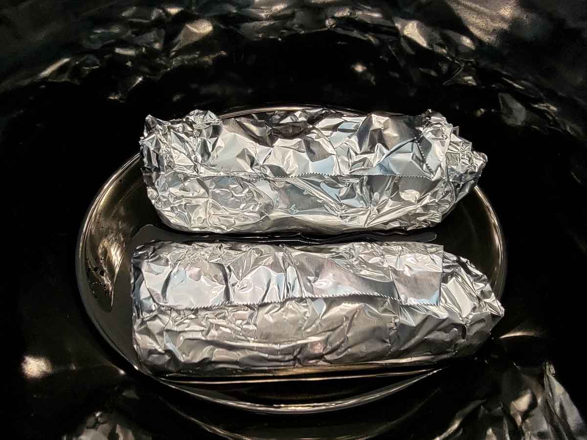 two foil wrapped corn cobs in a crock pot.