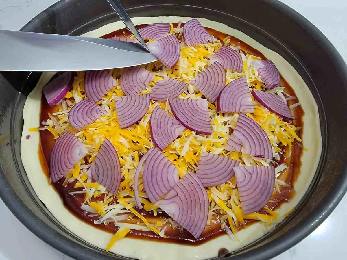 red onions slices and shredded cheese added to pizza dough and sauce in a pan.