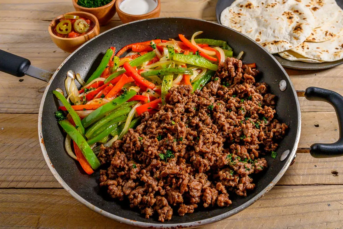 Ground Beef Fajitas in a skillet with sides of tortillas, salsa, sour cream, and parsley.