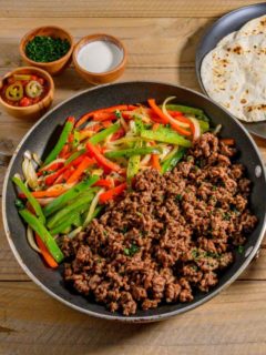 Fajitas with Ground Beef in a skillet with sides of tortillas, salsa, sour cream, and parsley.