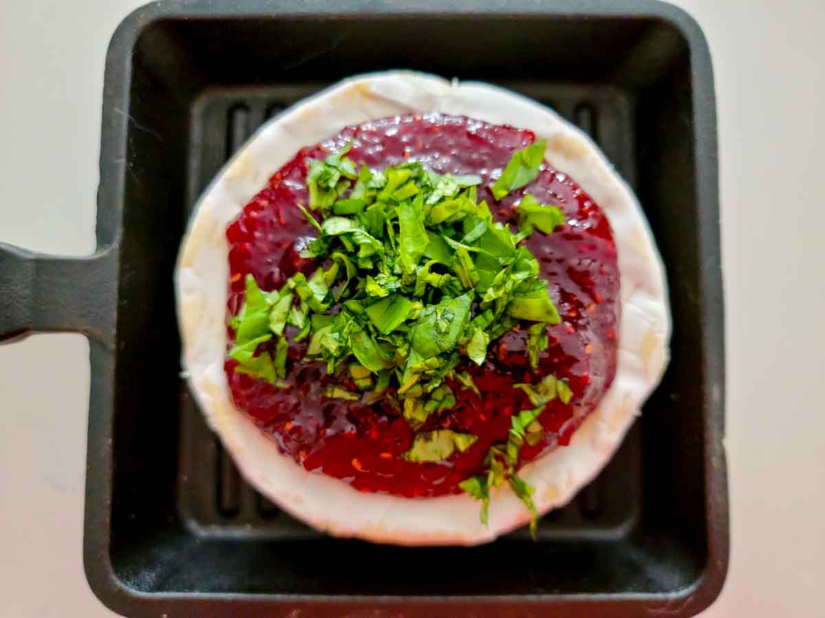a wheel of unbaked brie cheese with jam and basil on top in a small cast iron dish.