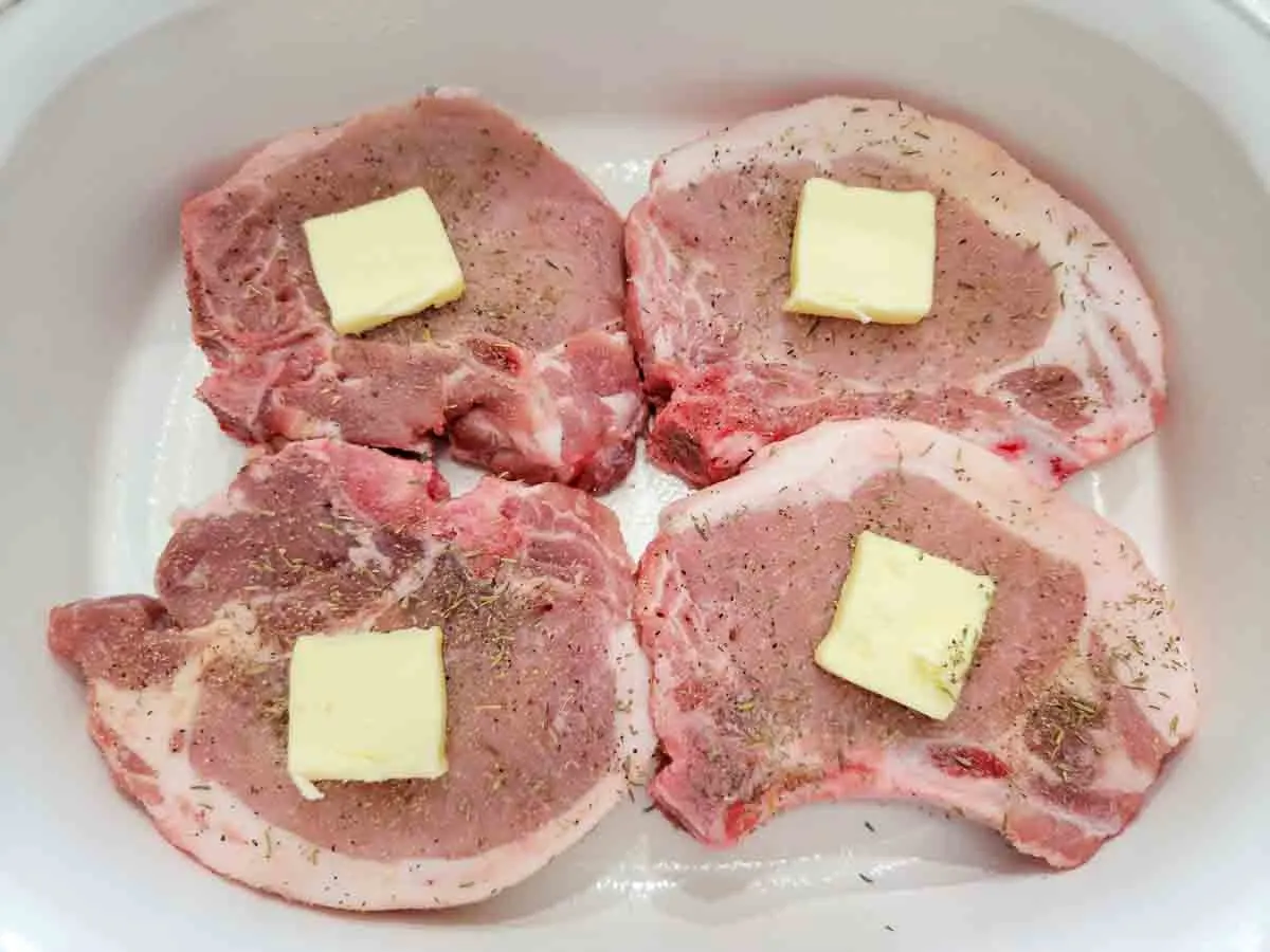 four pork chops topped with herbs, butter, and seasonings in a baking dish.