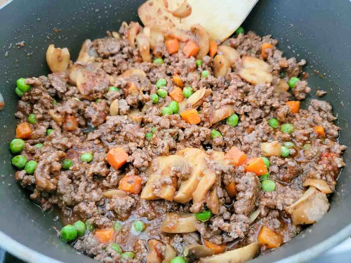 ground beef, mushrooms, peas, and carrots, cooking in a pan.
