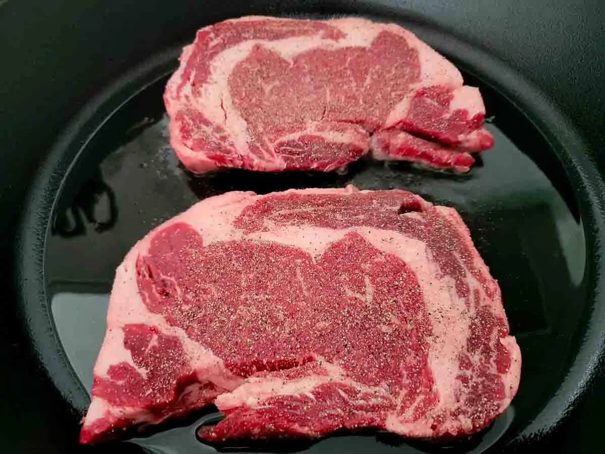 ribeye steaks evenly sprinkled with salt, pepper, garlic powder, and onion powder cooking in a cast iron skillet.