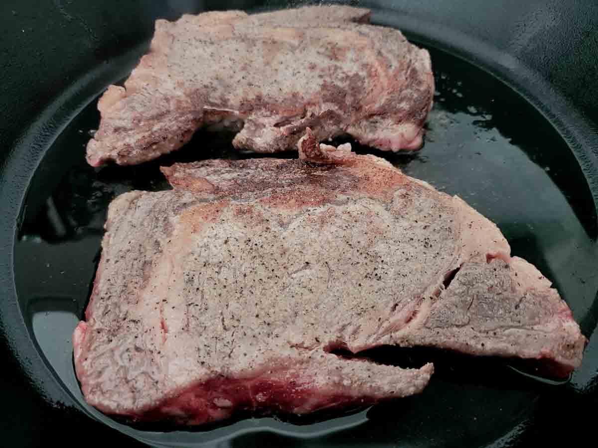 ribeye steaks evenly sprinkled with salt, pepper, garlic powder, and onion powder cooking in a cast iron skillet.