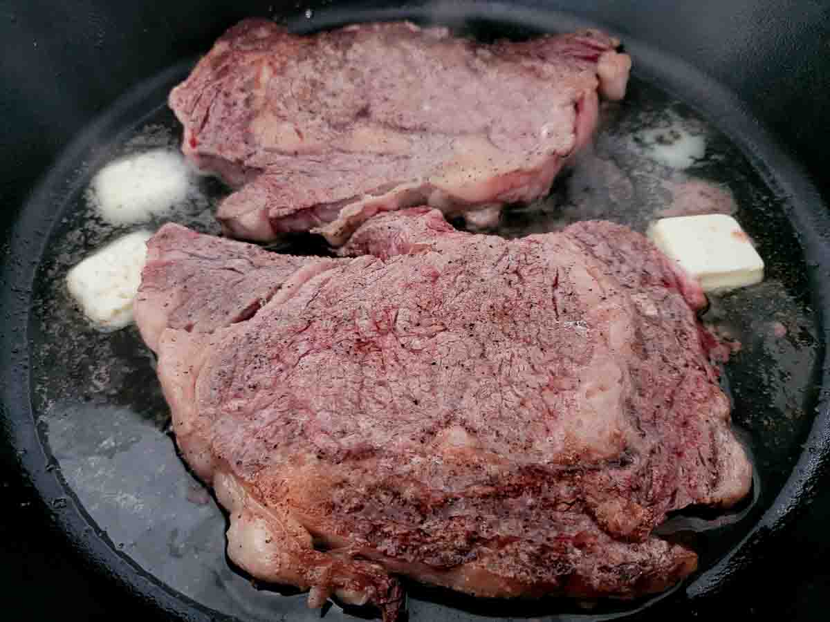 butter, ribeye steaks evenly sprinkled with salt, pepper, garlic powder, and onion powder cooking in a cast iron skillet.