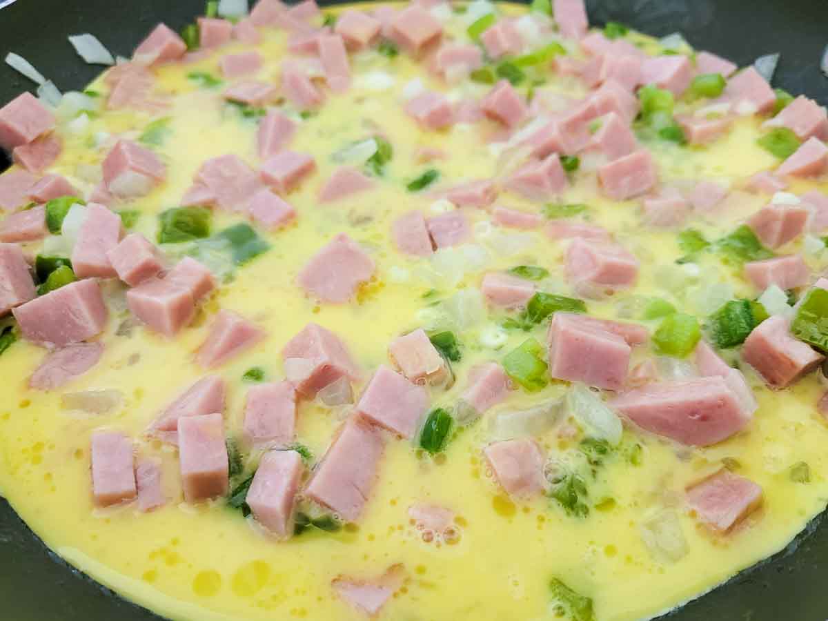 eggs, milk, diced spam, diced onions, and diced green peppers cooking in a skillet.