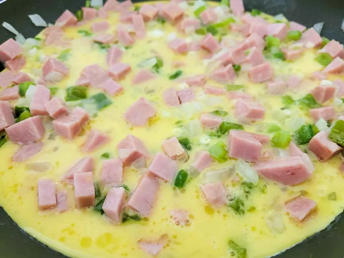 eggs, milk, diced spam, diced onions, and diced green peppers cooking in a skillet.