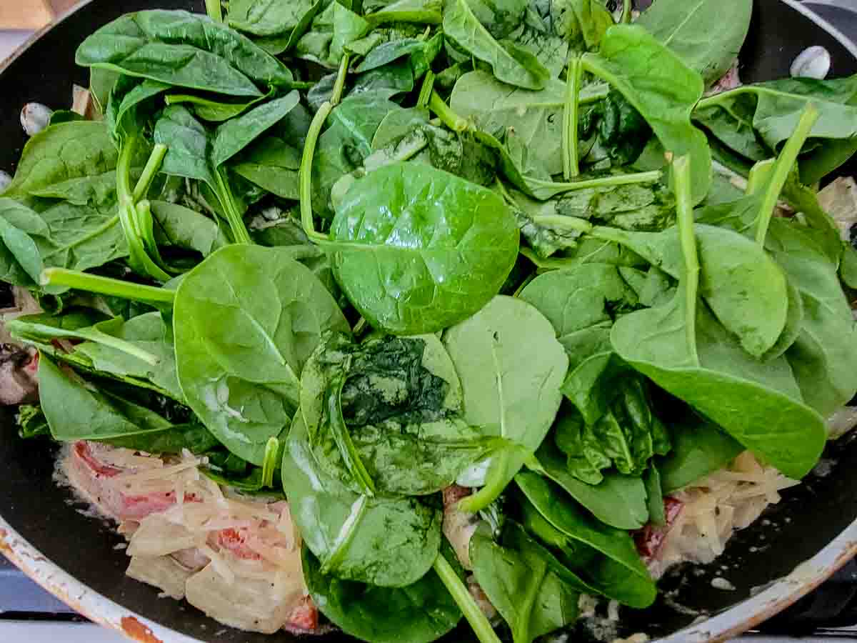 spinach, shredded parmesan cheese, heavy cream, chicken broth, butter, minced garlic, onions, mushrooms, and roasted red peppers cooking in a skillet.