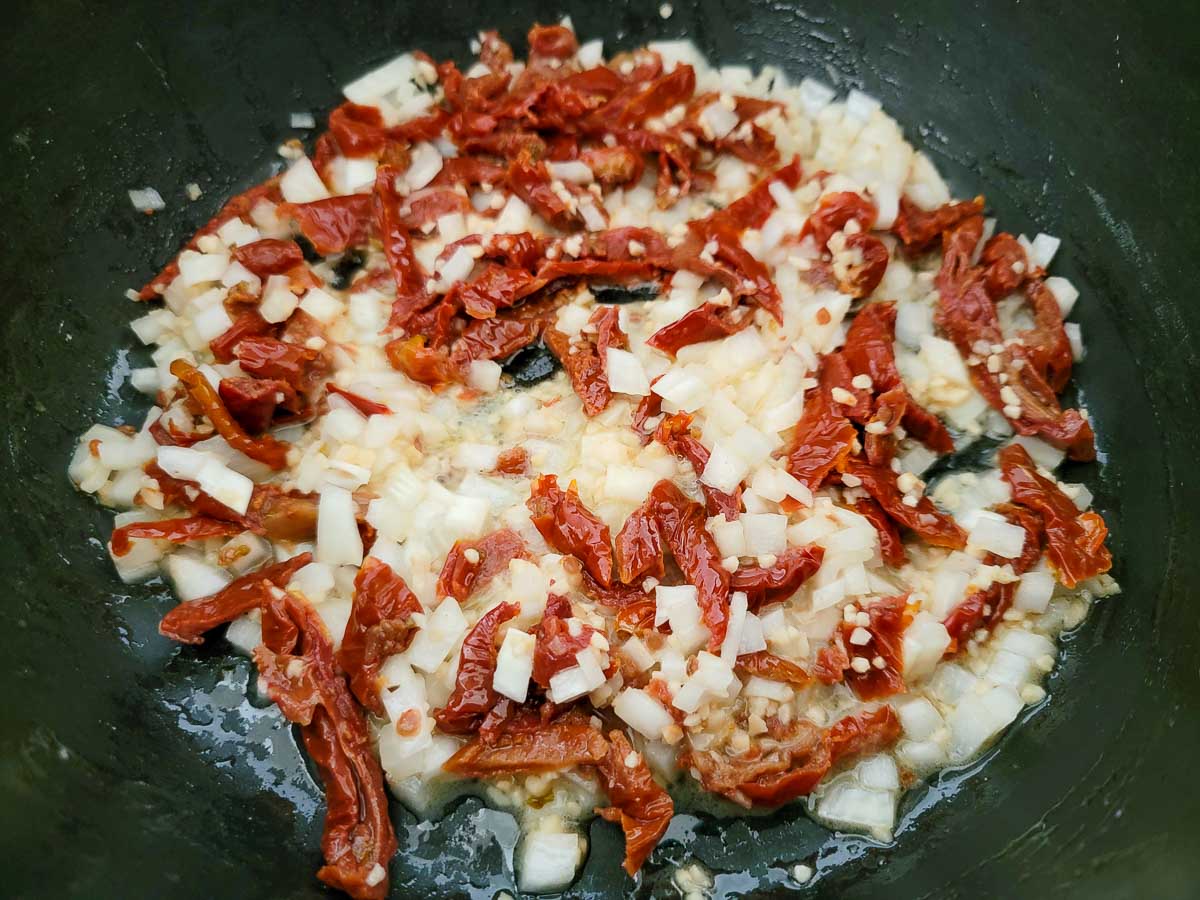 butter, minced garlic, onions, and sun-dried tomatoes cooking in a frying pan.