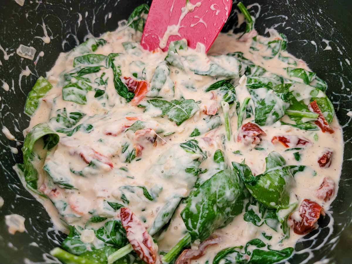 spinach leaves, Parmesan cheese, mozzarella cheese, heavy cream, butter, minced garlic, onions, and sun-dried tomatoes cooking in a frying pan.